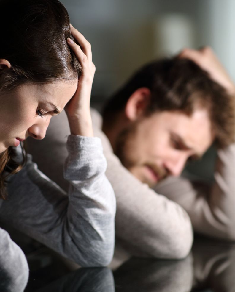 Couple leaning on a table holding their heads looking worried and sad as they are experiencing the pain of infidelity.