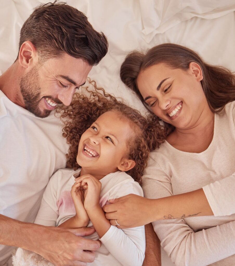Two parents with their daughter cuddling in bed looking happy representing the happiness you can feel after surviving infidelity. Committed couples can benefit from Infidelity Recovery Program by Relationship Experts Online serving couples in the Untied States, Canada, and worldwide. Schedule a consultation with us today. 