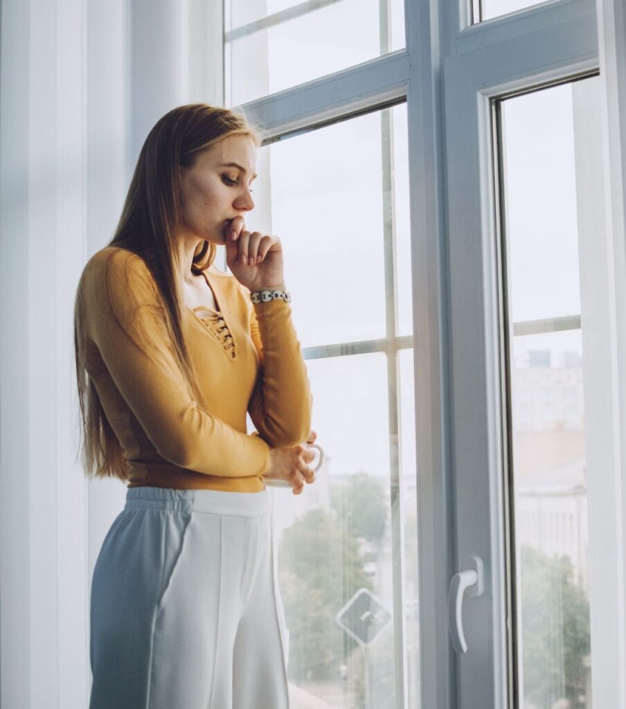 A woman standing next to a window, looking outside representing coping with PTSD and Infidelity. You can benefit from betrayal trauma recovery with Relationship Experts online. Schedule a consultation today.