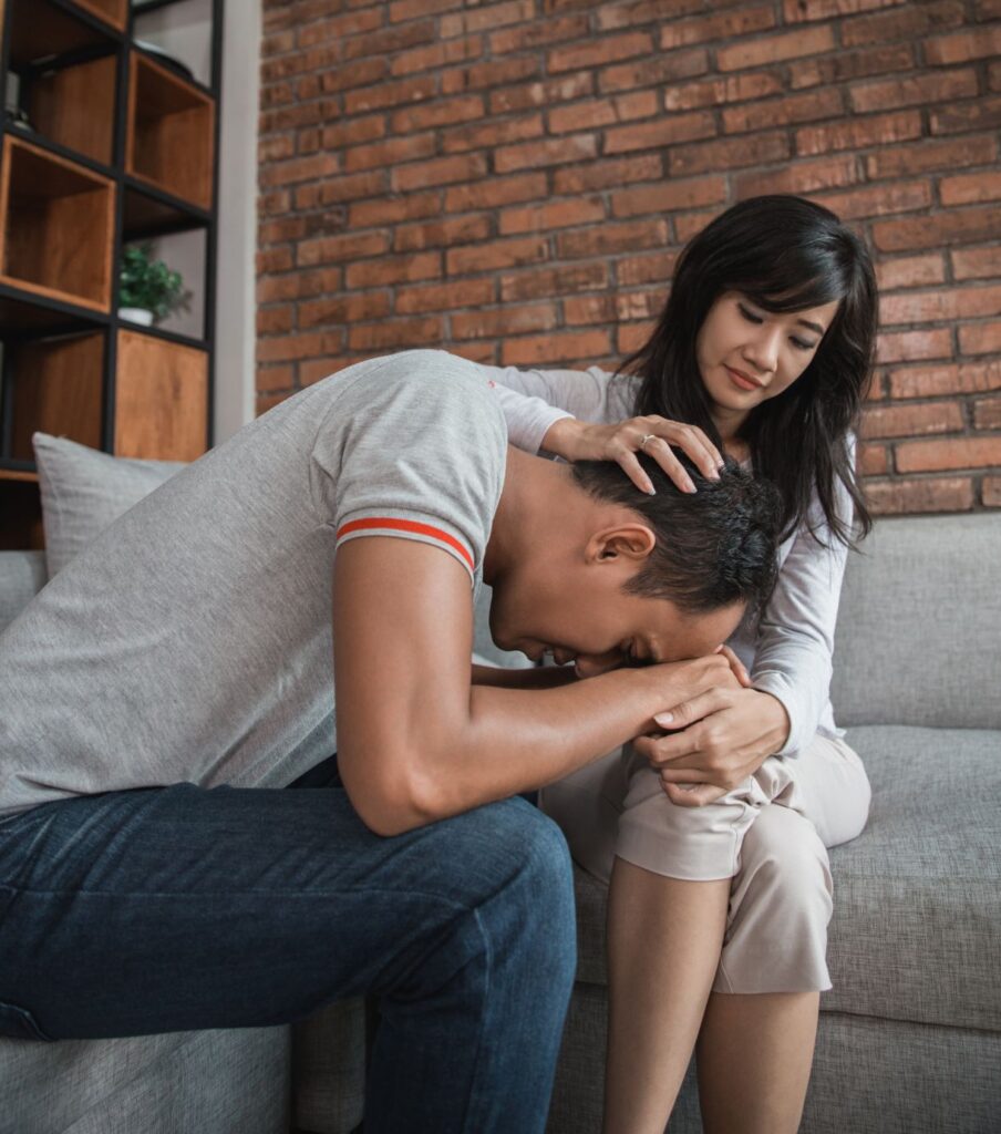 A man sitting with his head down while a woman puts her hand on his head to comfort him. This can represent a betraying partner trying to support her betrayed spouse. If you're the unfaithful partner and you want to know how to help your spouse heal from your affair, read more about our betrayal trauma recovery program in Florida, California, NY and the USA