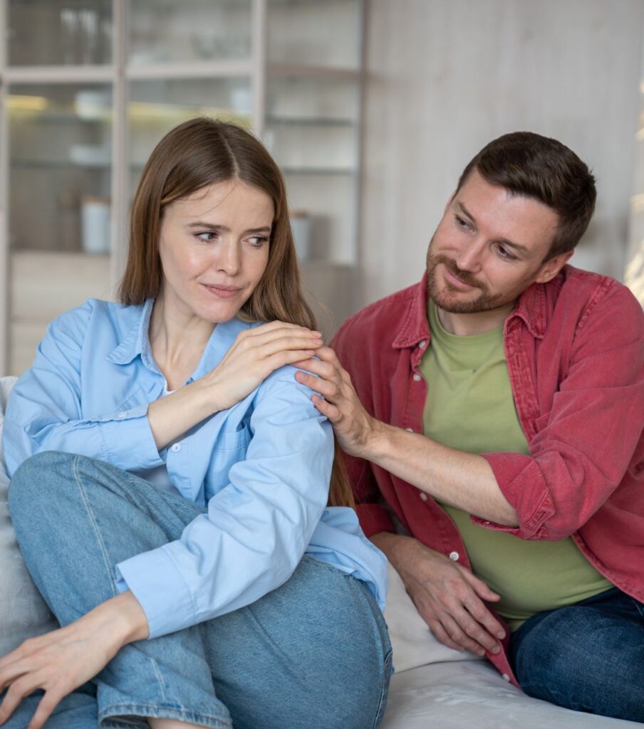 A smiling couple sitting on a sofa, with her touching his hand as he touches her shoulder, symbolizing the possibility to reconcile after cheating with Relationship Experts. Schedule a free consultation. Serving couples in North Carolina, the USA, Canada, the UK, and Australia.