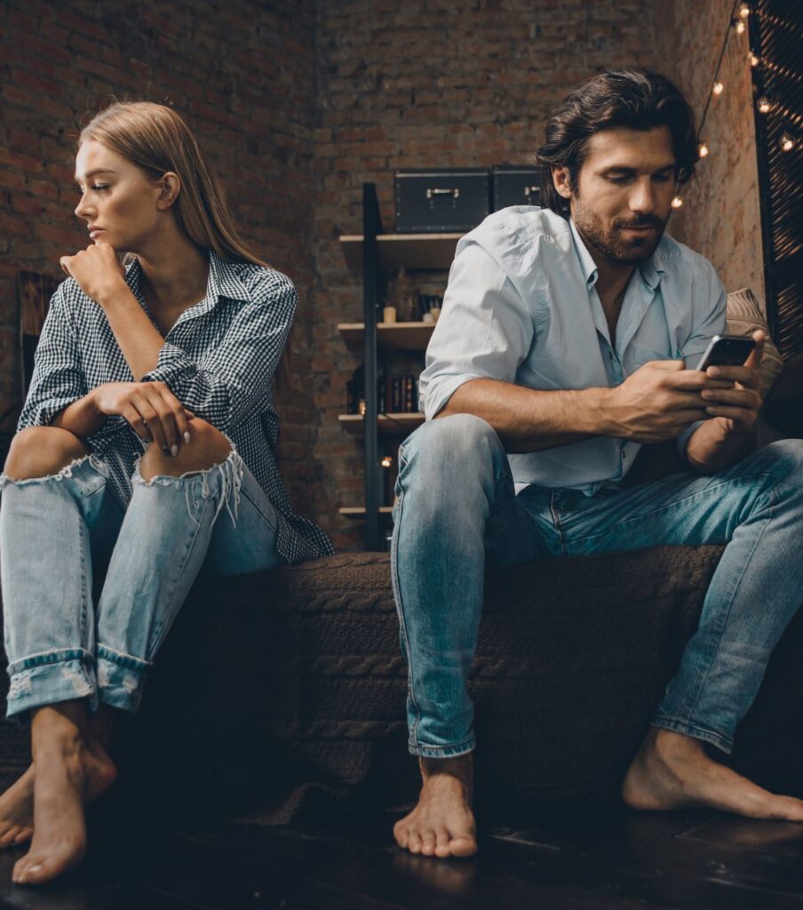 A couple sits on a bed with their backs turned, the man using his cell phone, symbolizing the struggle of reconciliation after infidelity. Get expert help to recover. Schedule a free consultation with a relationship expert in New York, in the United States, Canada, and the UK.