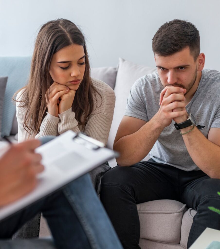 Couple in a therapist's office, sitting on a sofa with their faces in their fists, symbolizing the 'trickle truth' approach over transparency. Contact Relationship Experts in New York, California, and globally for healing after cheating.