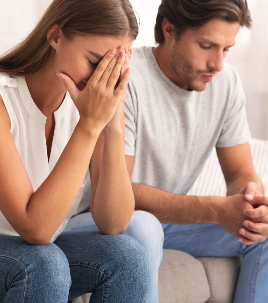 A man sitting beside a woman covering her eyes, symbolizing the emotional challenges couples face before healing. Infidelity recovery programs do help rebuild relationships after infidelity in the USA and beyond.
