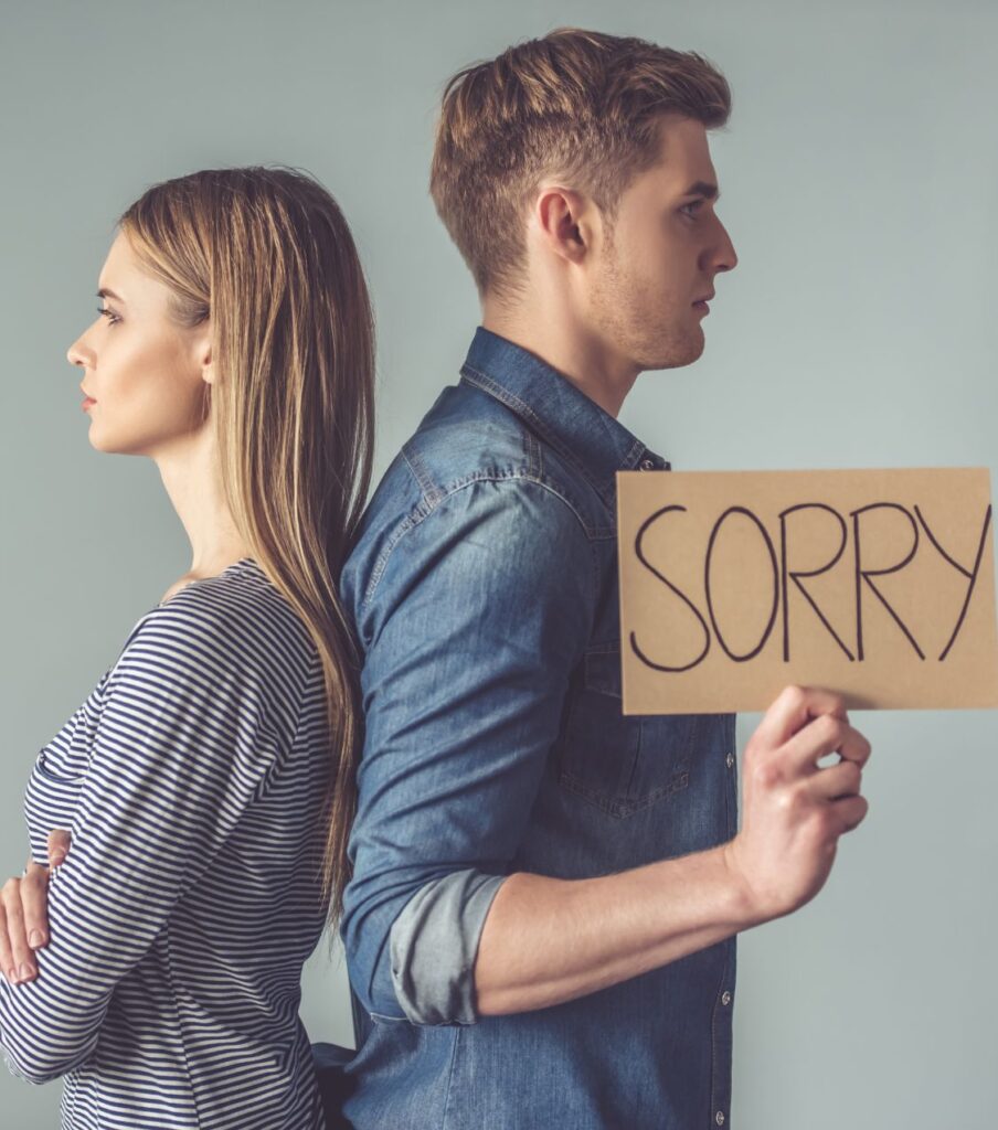 A man stands holding a "sorry" sign with his back to a woman, symbolizing showing remorse after cheating. Our therapists help couples heal after cheating with specific, intentional steps in the USA, Canada, the UK, and worldwide. Schedule a call today

