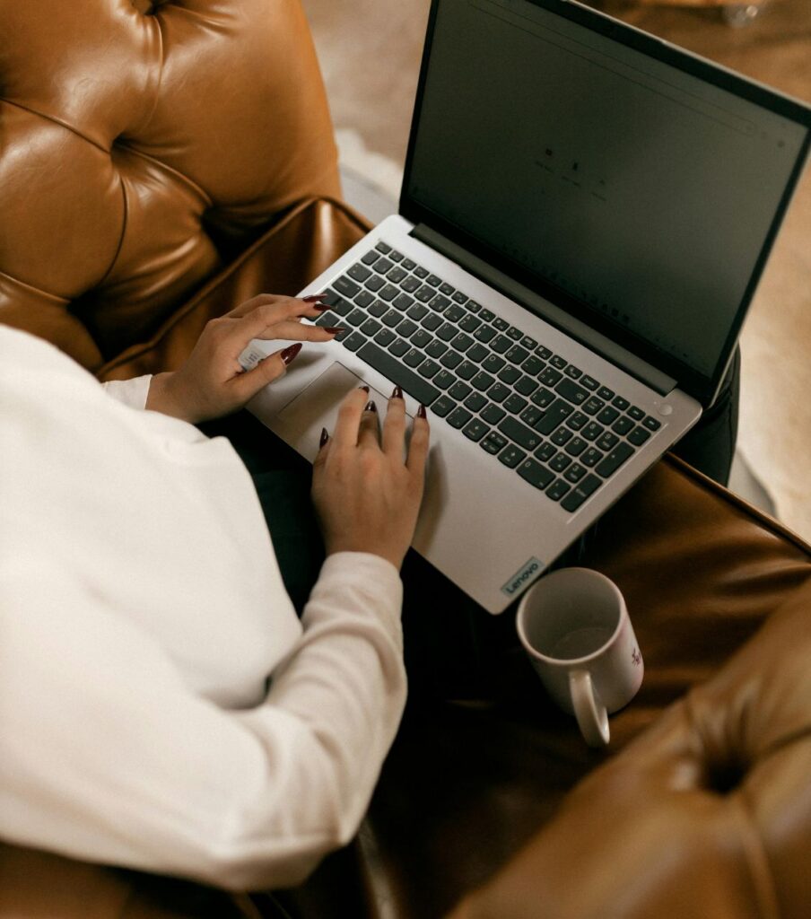 A woman types on a laptop while seated with a coffee cup nearby, representing someone researching expert help after a partner had a same-sex affair and realizing that same-sex betrayal recovery requires specialized dual training in affair recovery and sex therapy. Our licensed couples therapists serve clients in the USA, UK, and Canada online. Explore our affair recovery program and take the next step toward healing today.