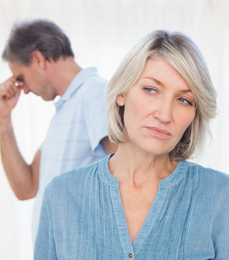 Woman in the foreground looking away with a hurt, unconvinced expression while a man stands behind her rubbing his forehead after a long apology. Illustrating why isn't apologizing enough to rebuild trust and how long does it take to rebuild trust after an affair. For couples working to rebuild trust after cheating in Australia, Dubai, the USA and globally. Explore our affair recovery program to learn what accountability looks like beyond an apology.