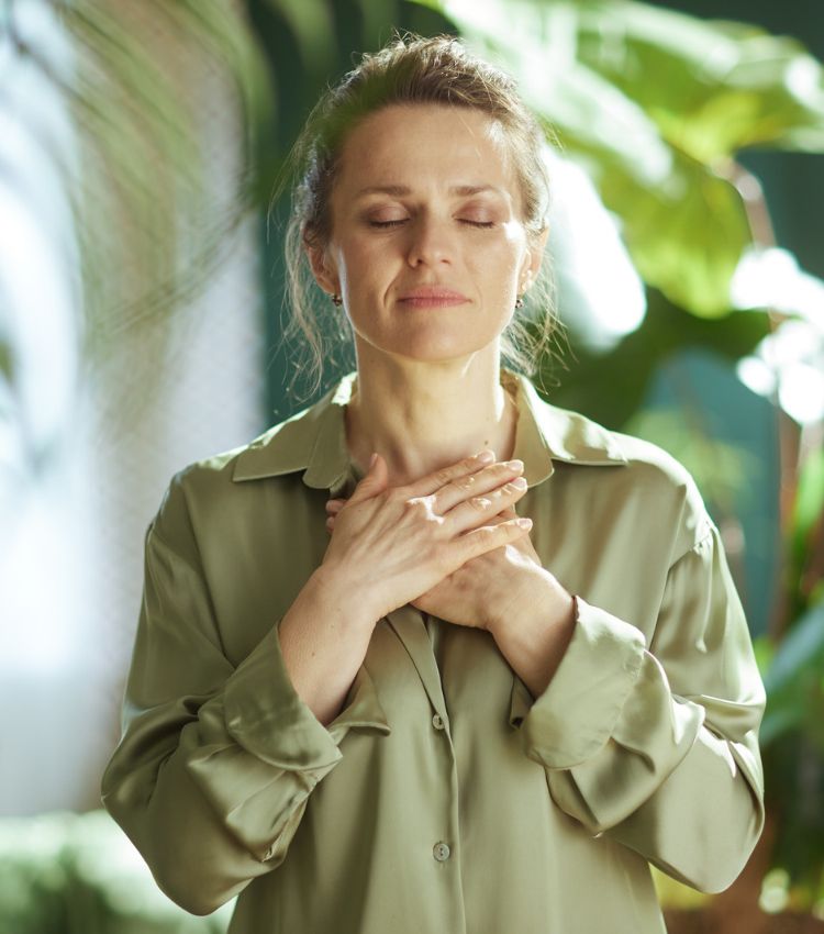 A woman with light brown hair pulled back, stands with her eyes closed and both hands crossed over her chest, surrounded by large leafy plants in soft light. In EMDR for infidelity, grounding and self-soothing tools come before bilateral stimulation, helping hurt partners manage betrayal trauma symptoms, with online support in Australia, Ireland, and New Zealand. Talk with our team to learn whether EMDR is the right next step.